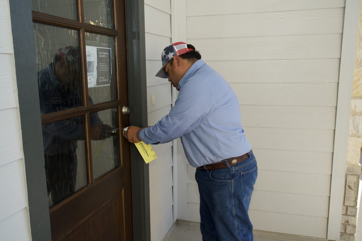 Man in cap and light blue shirt attaching a yellow notice to a glass paneled front door