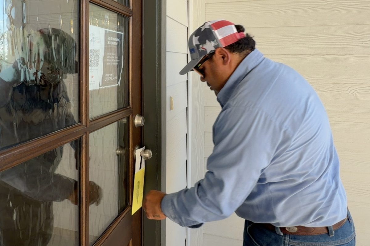 Man in cap and blue shirt attaching a yellow door hanger to a glass paneled front door