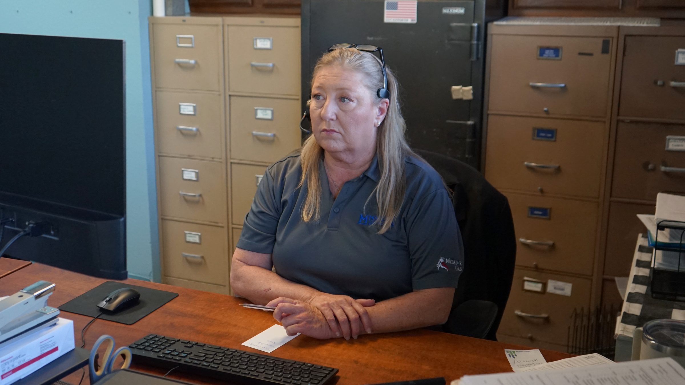 Woman wearing a headset sits at an office desk with filing cabinets and a safe in the background