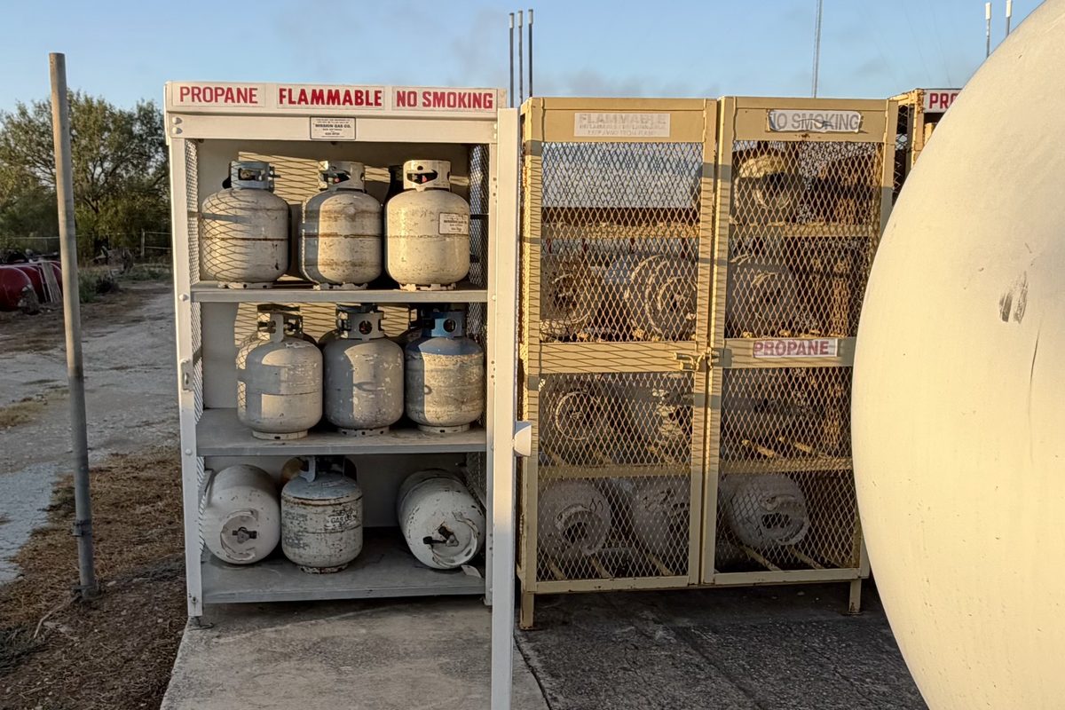 Propane cylinder exchange cages with tanks on shelves beside a large storage tank