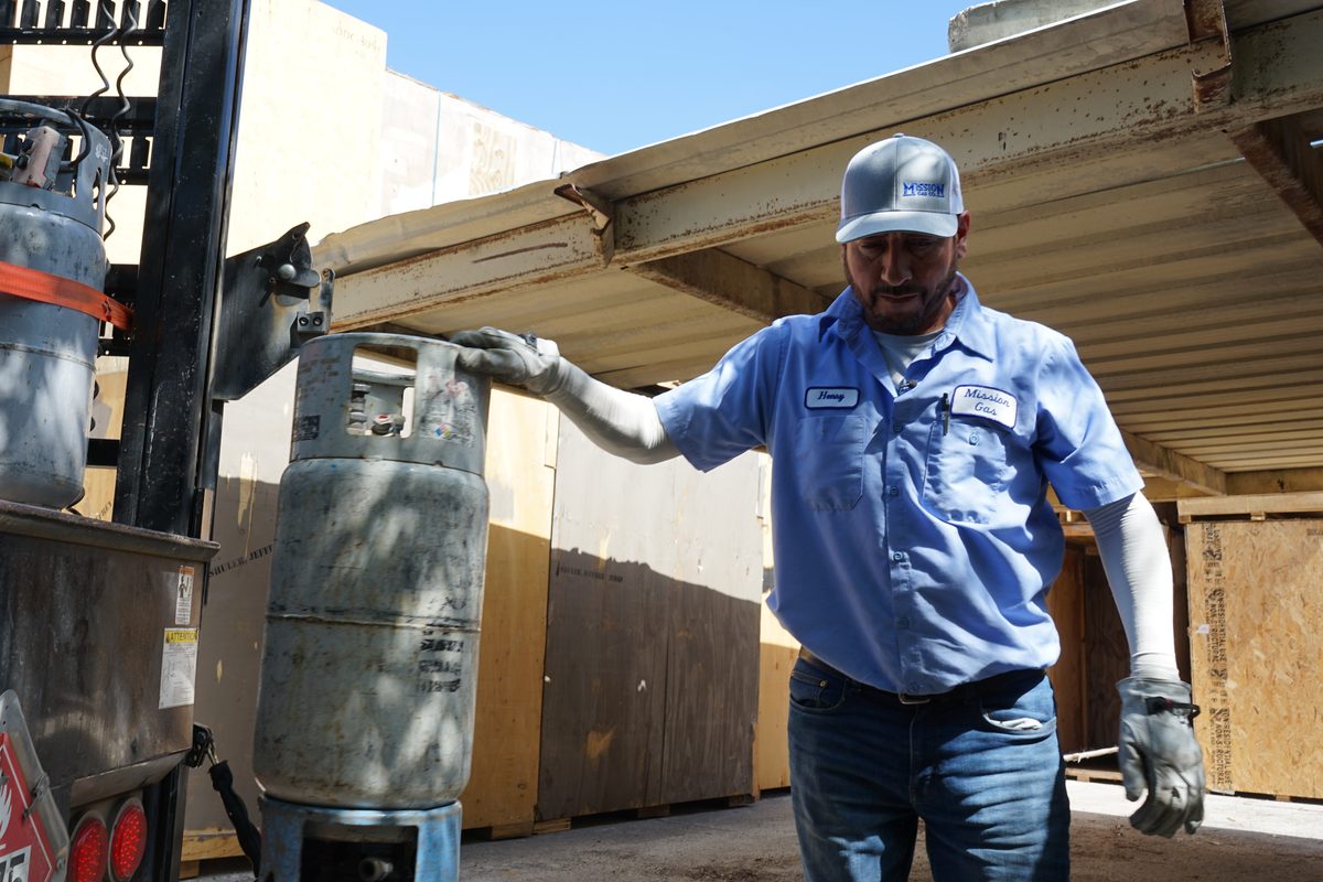 Worker in Mission Gas shirt guiding a propane cylinder off a forklift at a storage yard