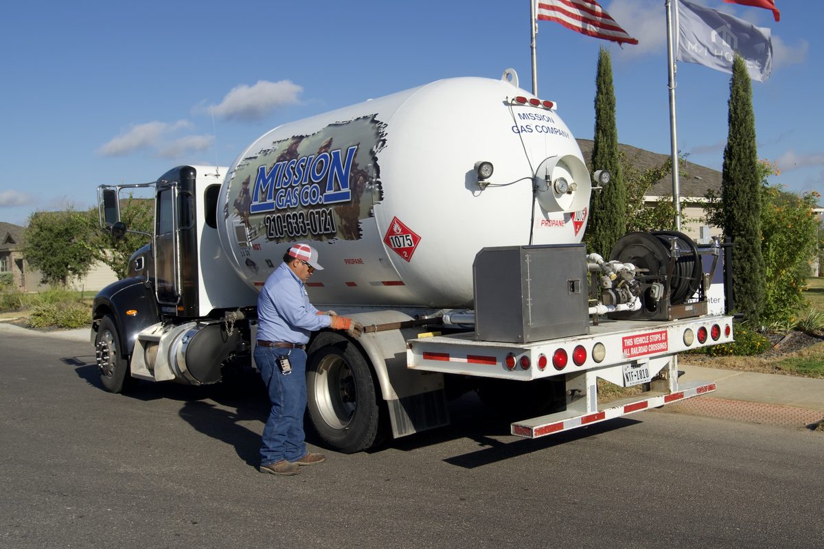Mission Gas Co. propane truck parked on a street with a worker checking rear equipment, American and Texas flags flying