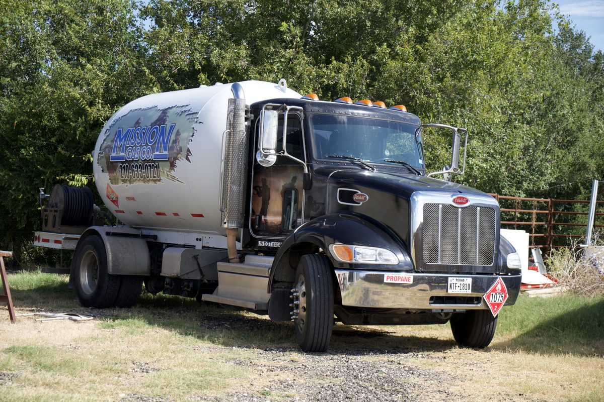 Black Peterbilt propane delivery truck with white tank labeled Mission Gas Co. parked on gravel near trees
