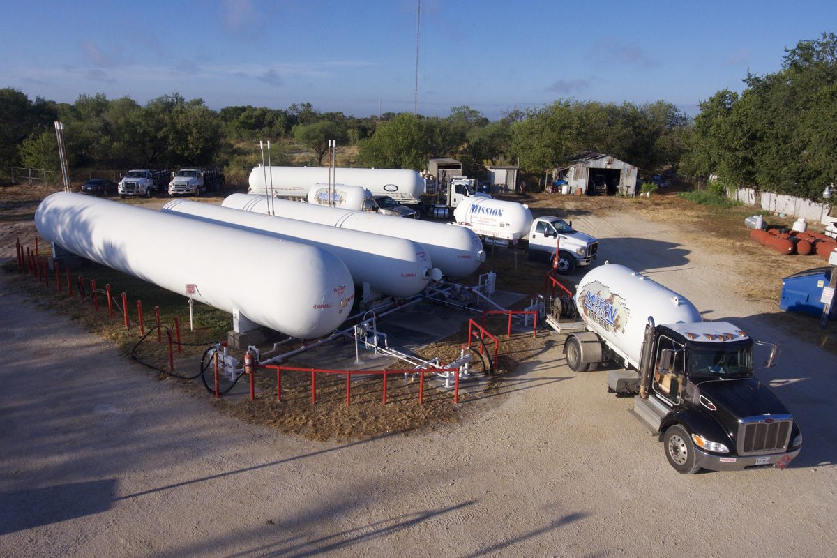 Large propane storage tanks and a Mission Propane delivery truck parked in a rural yard with trees.