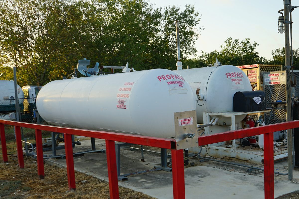 White propane storage tanks with warning labels inside a red metal safety fence at a fuel yard