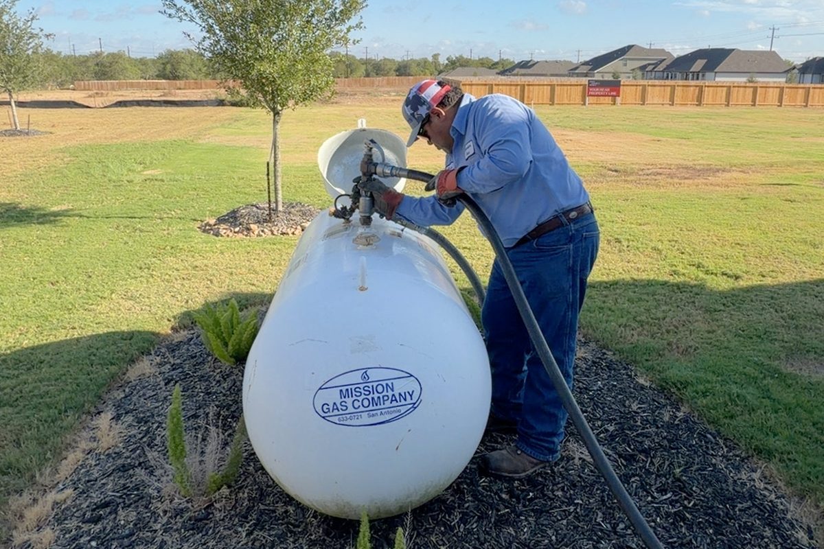 Technician in blue shirt filling a residential propane tank labeled Mission Gas Company on a landscaped mulch bed