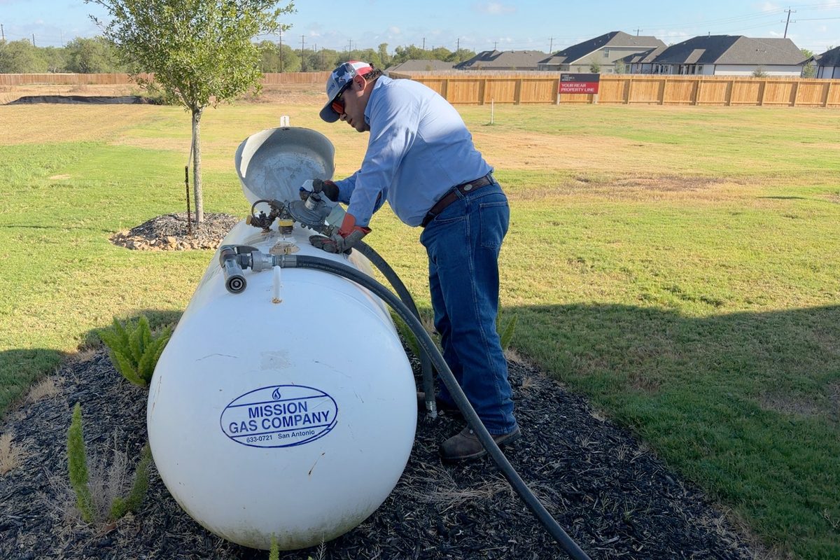 Technician in cap and blue shirt servicing a white Mission Gas Company propane tank on a grassy lot