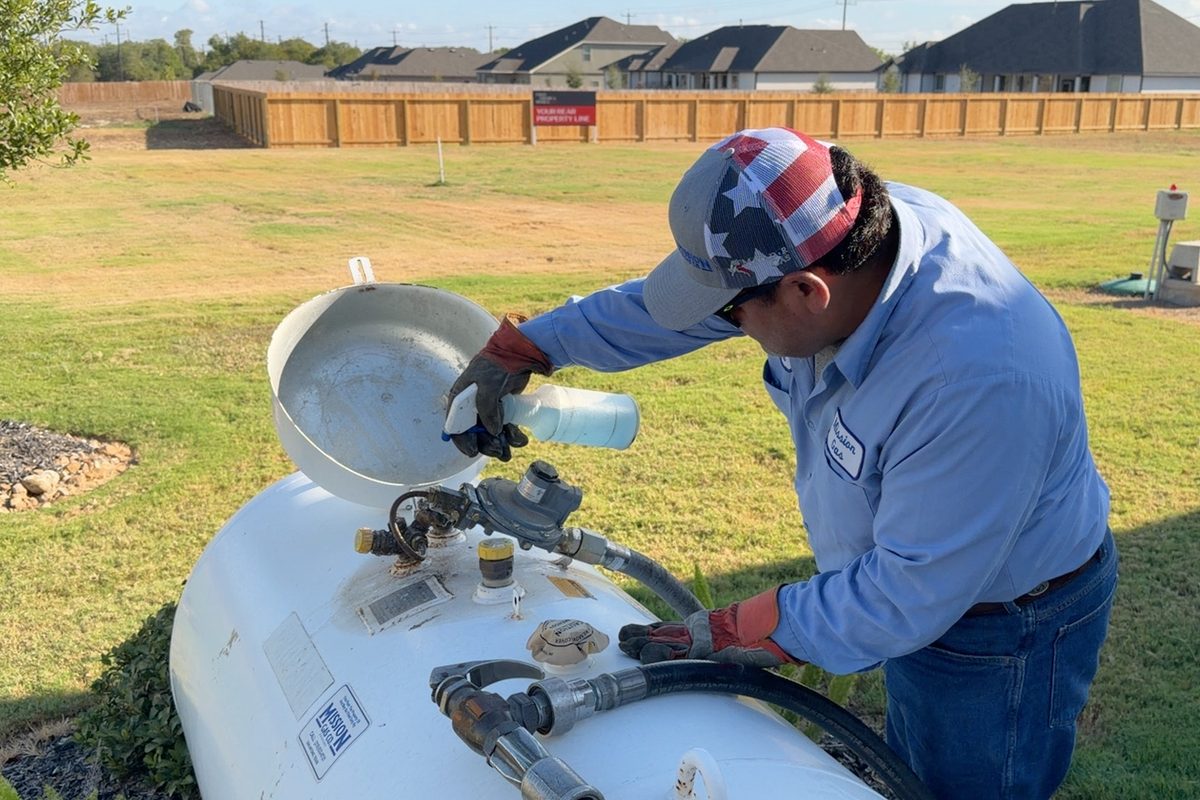 Technician in cap and work shirt services a white propane tank in a residential yard