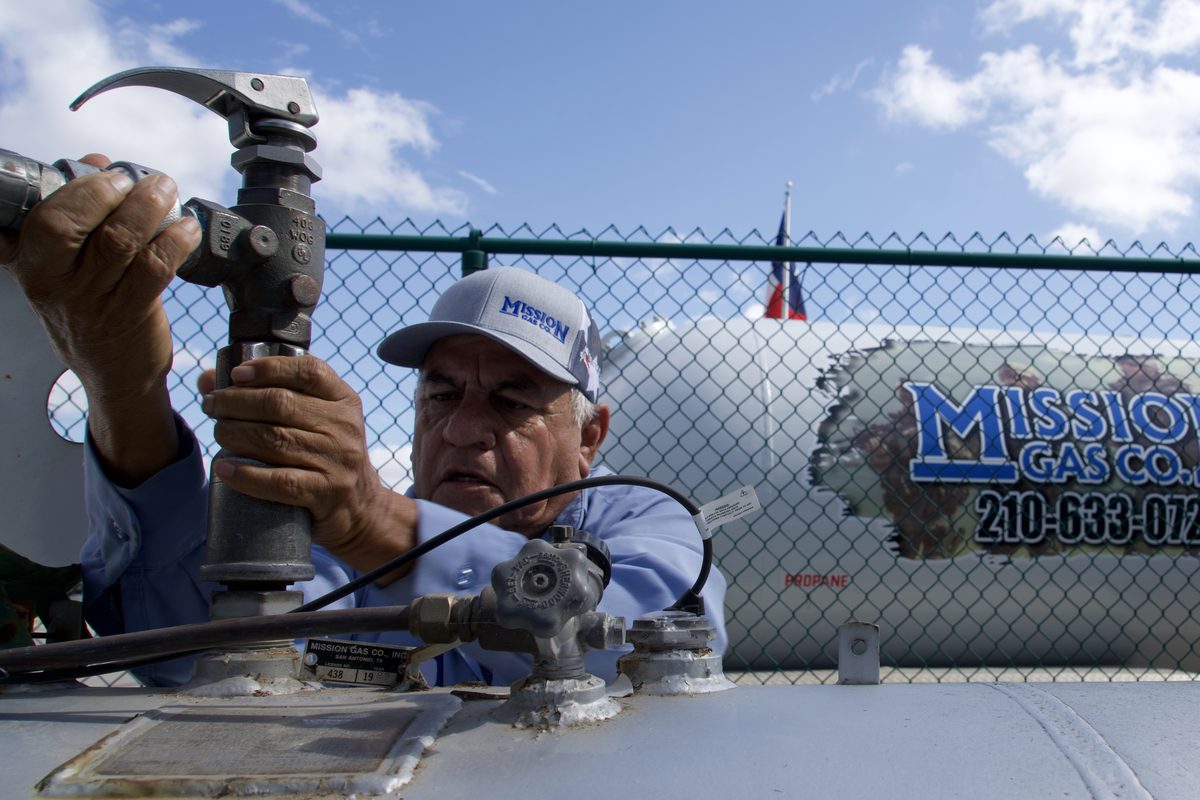 Man in Mission Gas Co cap adjusting a valve on a propane tank with company signage in background