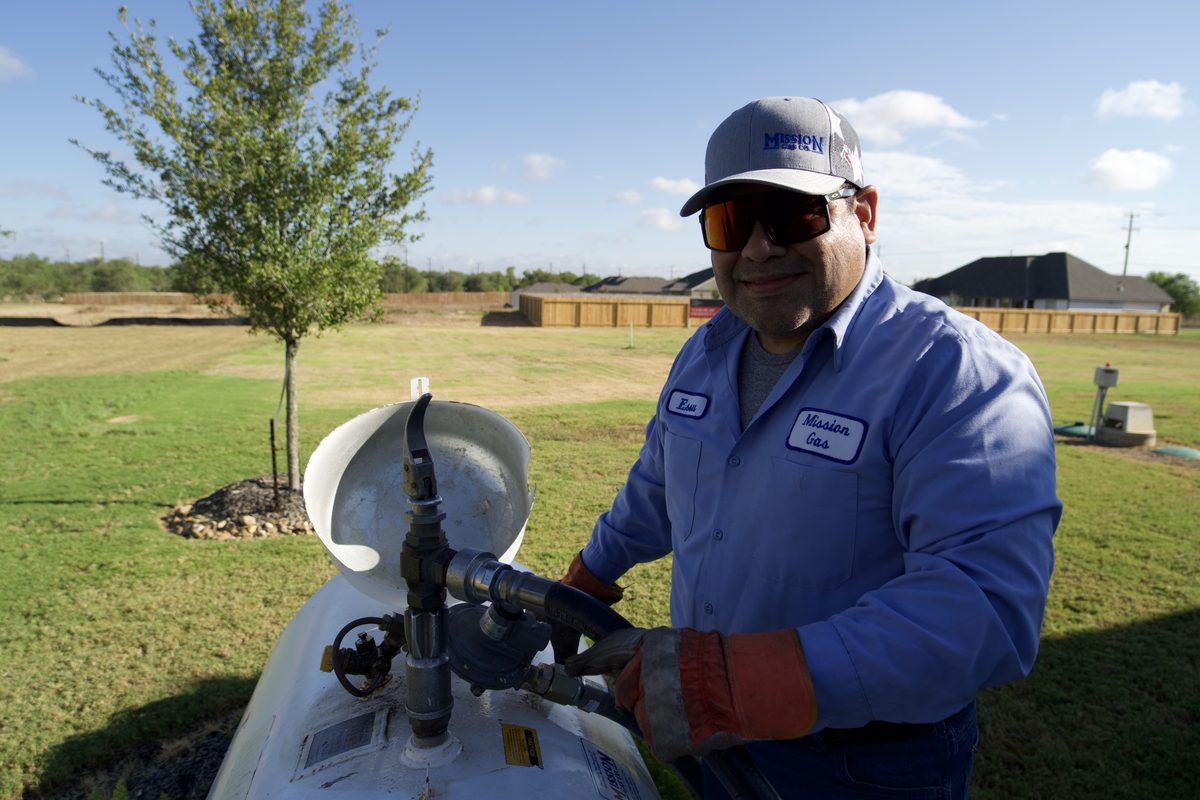 Technician in a blue Mission Gas uniform servicing a propane tank's valve outdoors