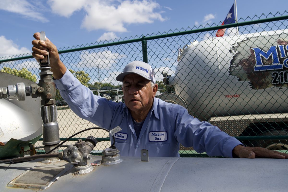 Technician in Mission Gas uniform adjusts valve on propane tank, with Mission Gas Co. truck behind chain link fence