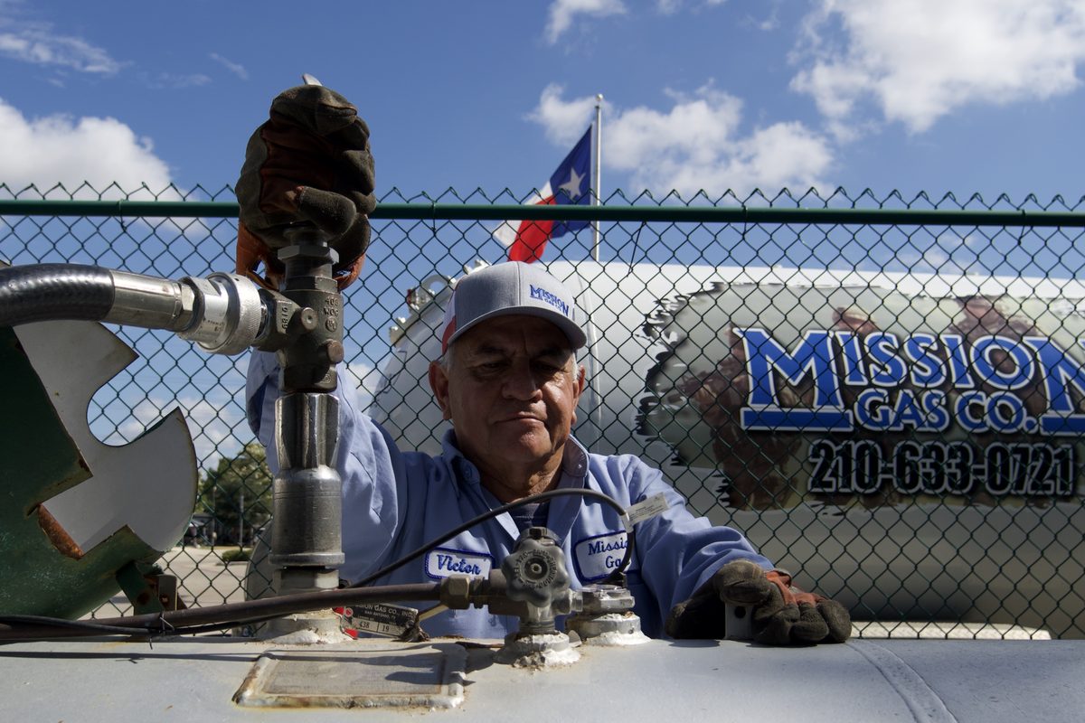 Technician in Mission Gas Co uniform opens a valve on top of a propane tank under a blue sky