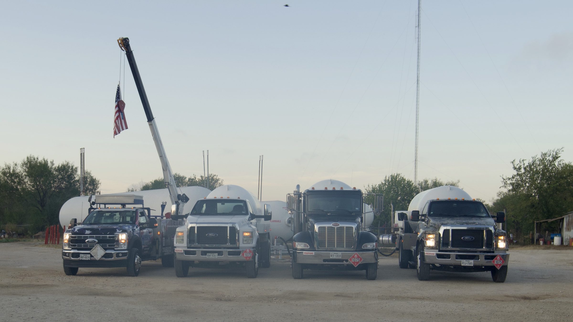 Fleet of propane delivery trucks and a crane truck parked in a gravel yard at dusk