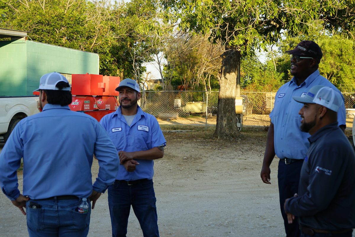 Four propane delivery workers in uniform shirts and caps talking outdoors near a truck