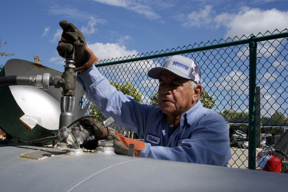 Worker in a blue shirt and cap adjusts a valve on a propane tank against a fenced lot and blue sky