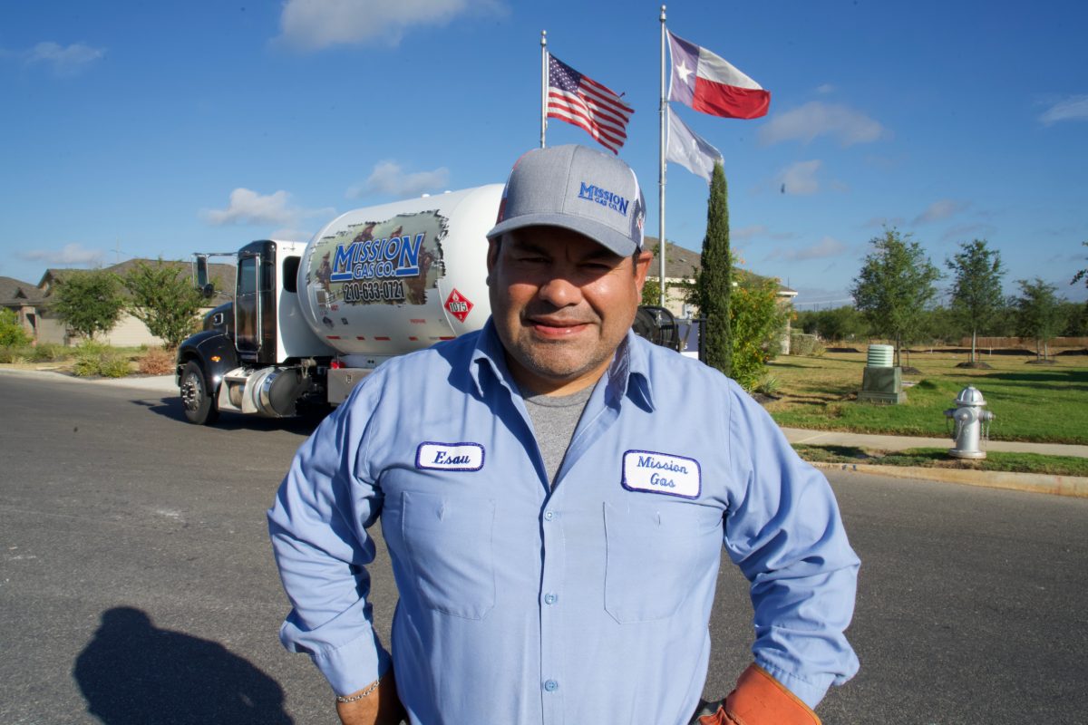 Smiling Mission Gas delivery driver in uniform standing in front of a propane truck with American and Texas flags behind him