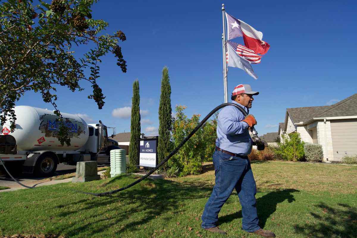 Mission Gas driver in a cap pulling a delivery hose across a front lawn from a propane truck