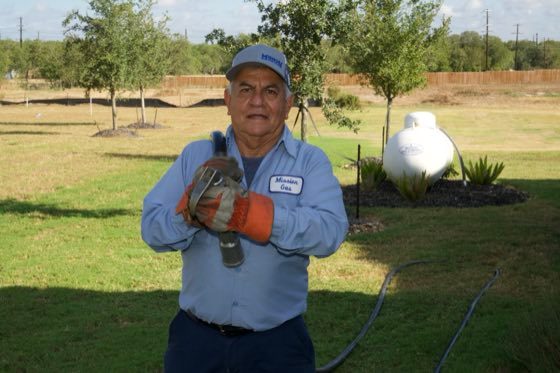 Mission Gas technician in work gloves holding a propane hose in a yard with a white tank behind him